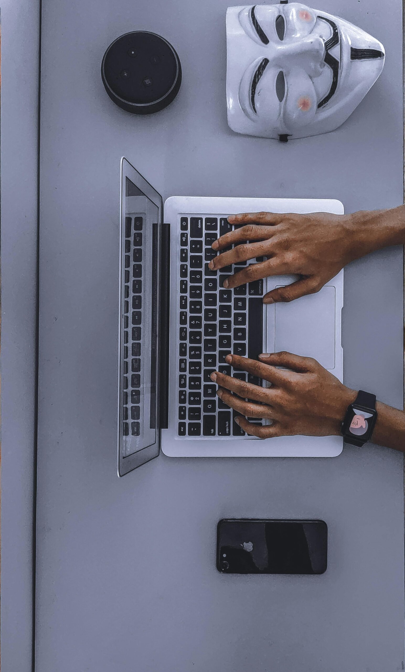 Top view of hands typing on a laptop beside a mask and smartphone.